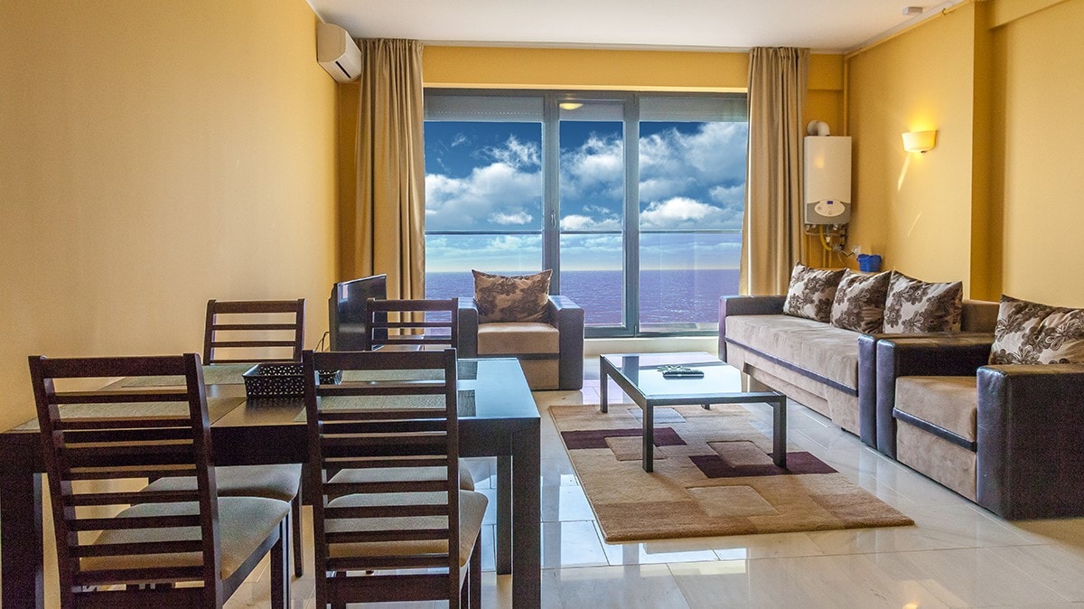 A spacious living area is presented with soft neutral tones and large windows displaying a view of the sea. Comfortable seating is arranged around a coffee table, while a dining table with chairs provides an inviting space for meals. Natural light fills the room.
