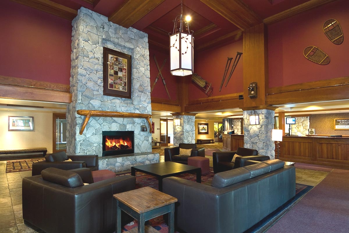 The lobby features high wooden beams and a large stone fireplace, creating a cozy gathering space. Leather sofas are arranged around a coffee table, with artwork and snowshoes displayed on the walls. Soft lighting enhances the welcoming atmosphere for guests.
