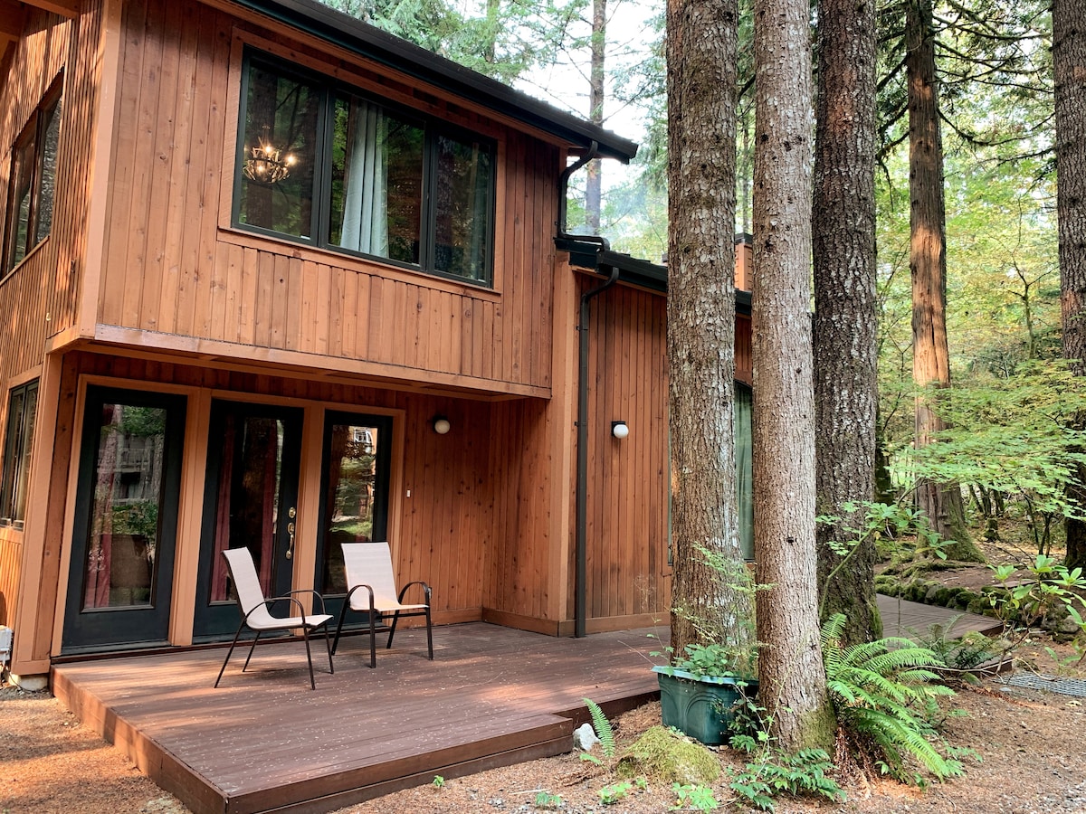 A wooden exterior of the lodge is framed by towering trees, creating a serene environment. The deck features two light-colored chairs facing the forest. Large windows offer glimpses into the cozy interior, emphasizing the connection between indoor and outdoor spaces.