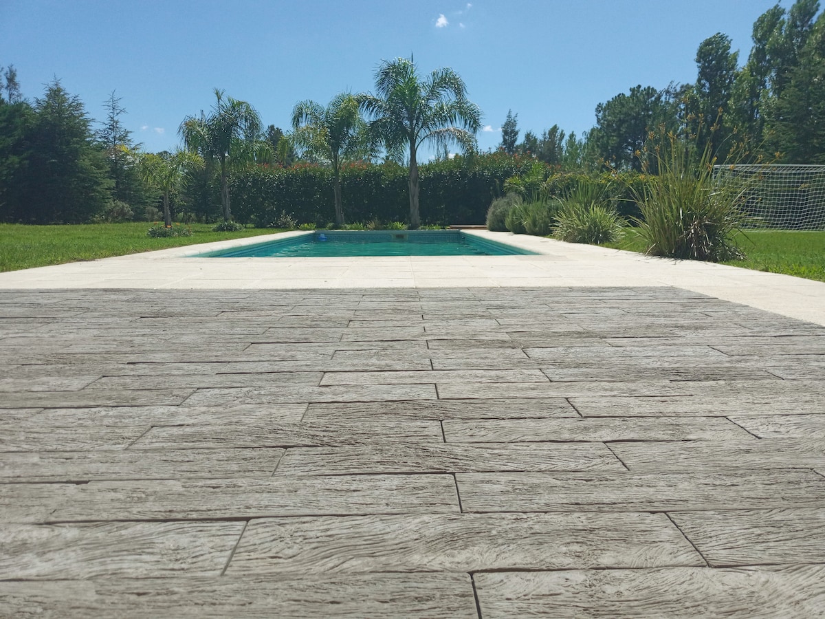 The image displays a serene swimming pool bordered by palm trees and lush greenery. The pool's clear water reflects the blue sky, while a textured wooden deck in the foreground adds a natural element. An expansive lawn surrounds the area, enhancing the tranquil atmosphere.