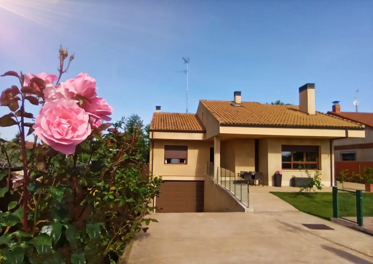 The exterior of the house is showcased, featuring a well-maintained facade with a charming porch and a sloped roof. Vibrant pink roses are in the foreground, and a well-manicured lawn surrounds the property, enhancing the inviting and private atmosphere.