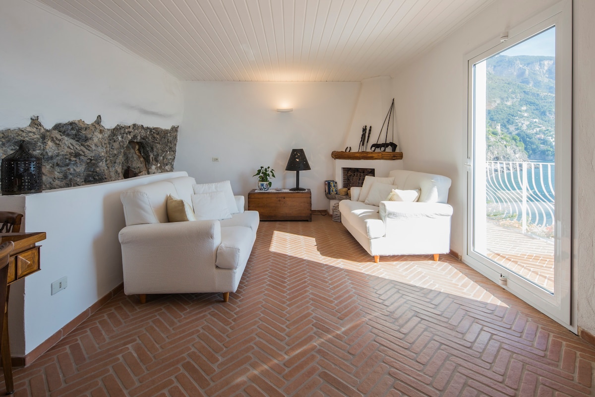 A bright living area features two white sofas positioned around a wooden coffee table. A textured stone wall adds character, while a large window offers views of the outside. Natural light fills the room, highlighting the warm terracotta flooring.