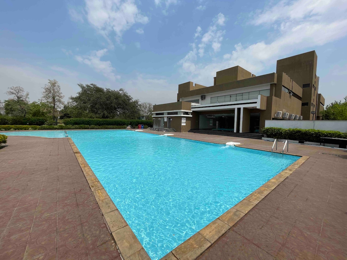 A spacious outdoor swimming pool is surrounded by a neatly maintained patio area, with sun loungers positioned along the water's edge. The building in the background houses a large terrace and multiple air conditioning units, set against a clear blue sky.