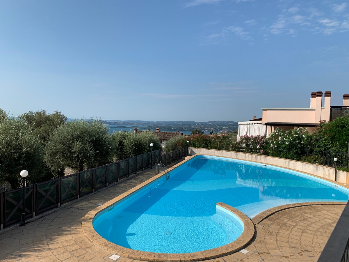 A serene swimming pool is shown, featuring clear blue water and a gently curved shape. Surrounding greenery, including olive trees and flowering plants, adds to the natural setting. A panoramic view of the lake and hills is visible in the background under a clear sky.