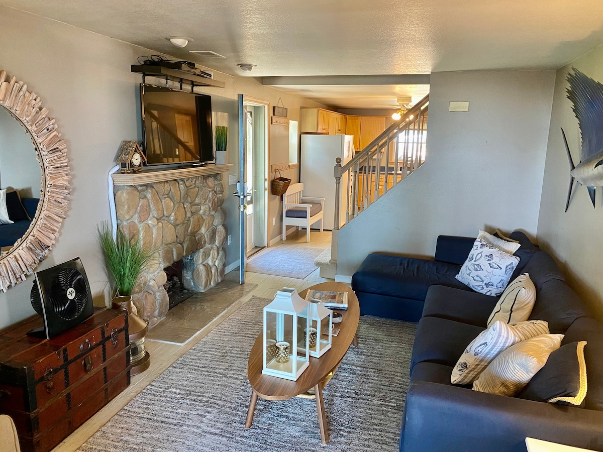 A cozy living area is furnished with a dark sectional sofa and a wooden coffee table. A stone fireplace is featured against the wall, complemented by a decorative mirror and nautical-themed accents. Light streams through the open space leading to the kitchen area.