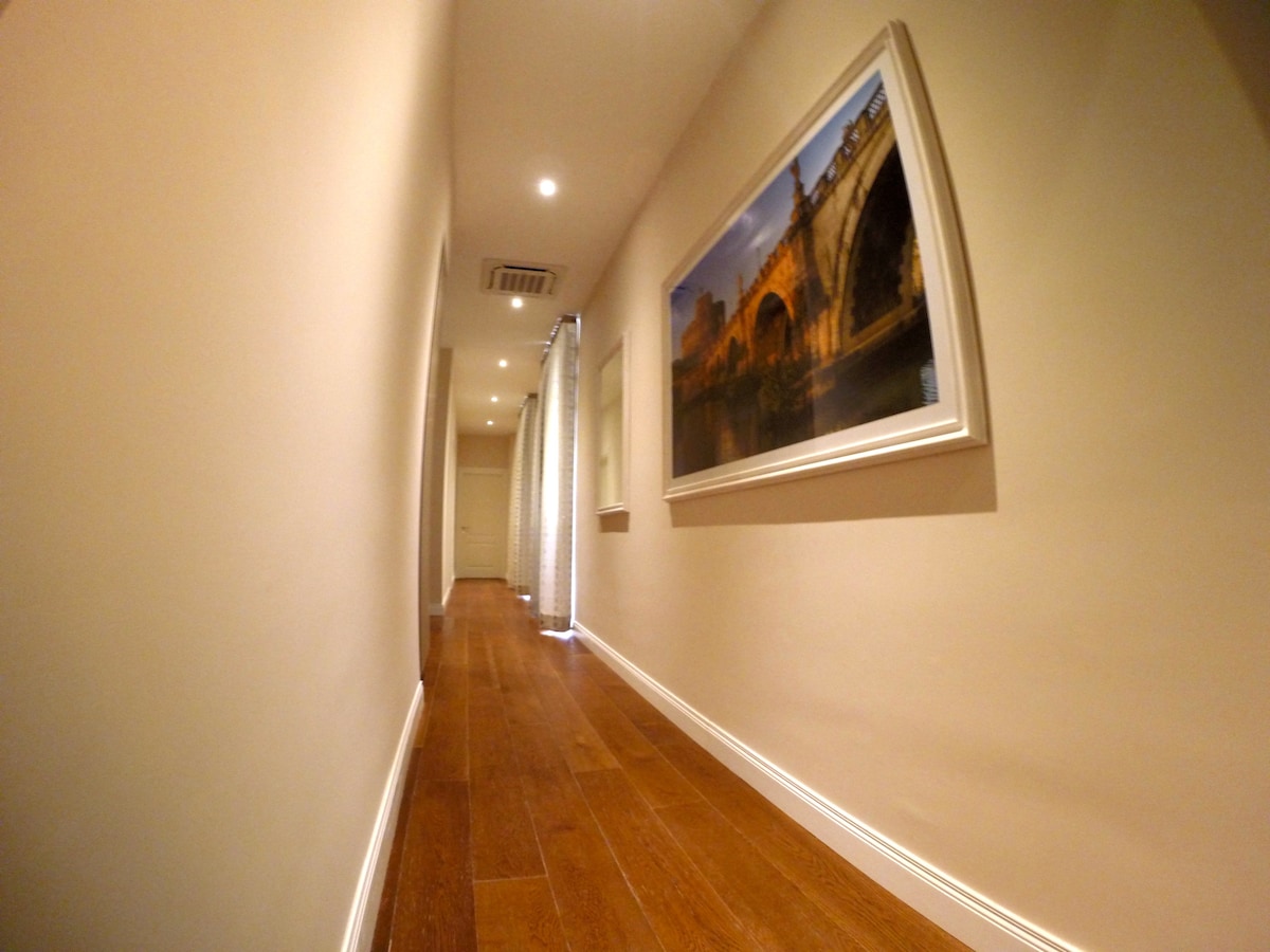 A long hallway featuring smooth wooden flooring, illuminated by recessed lighting. On the right, a large framed photograph captures an architectural scene, while light enters through a partially drawn curtain at the end of the hallway.