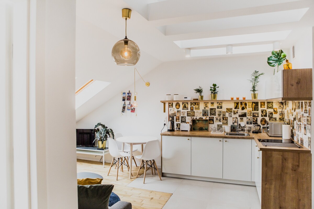 The kitchen area is bright and inviting, featuring a modern design with white cabinetry and wooden accents. A gallery of photographs decorates the wall above the counter. A round dining table with white chairs is positioned near a window, while greenery adds freshness to the space.