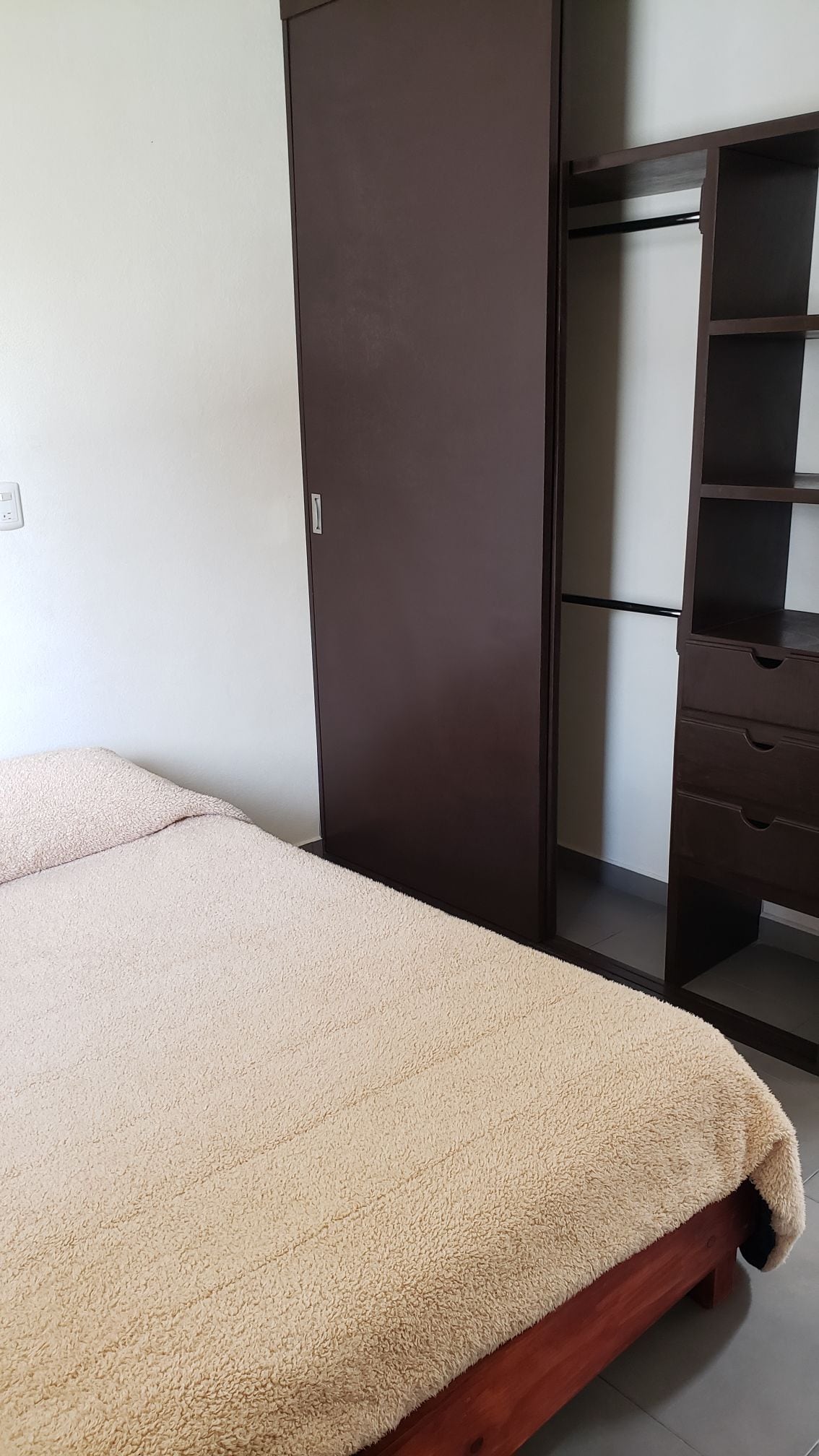 A simple sleeping area features a spacious bed covered with a soft beige blanket. A tall wardrobe and a shelving unit with drawers stand against the wall, providing storage options for personal belongings.