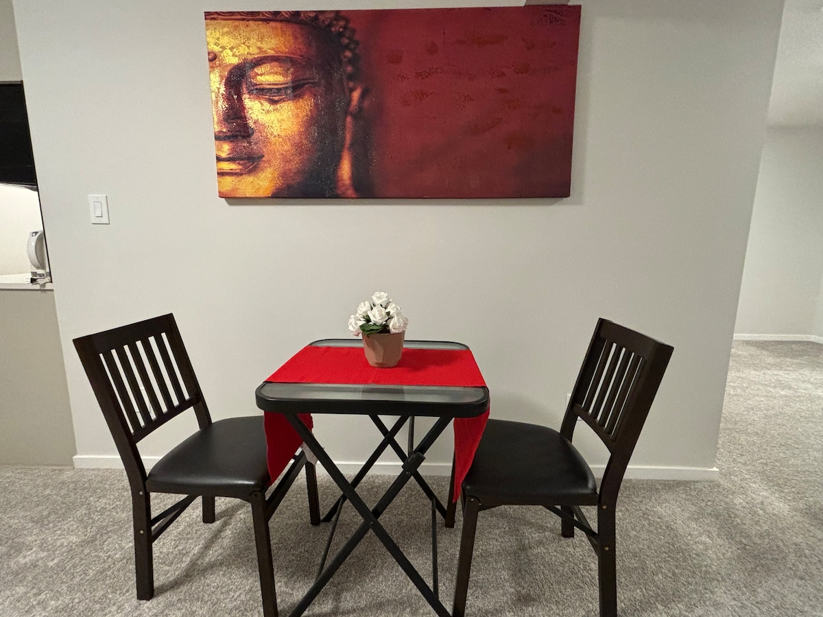 A compact dining area is featured, equipped with a small table and two wooden chairs. A red tablecloth is positioned atop the table, which holds a potted plant. A framed artwork is displayed above the table, adding an artistic touch to the space.