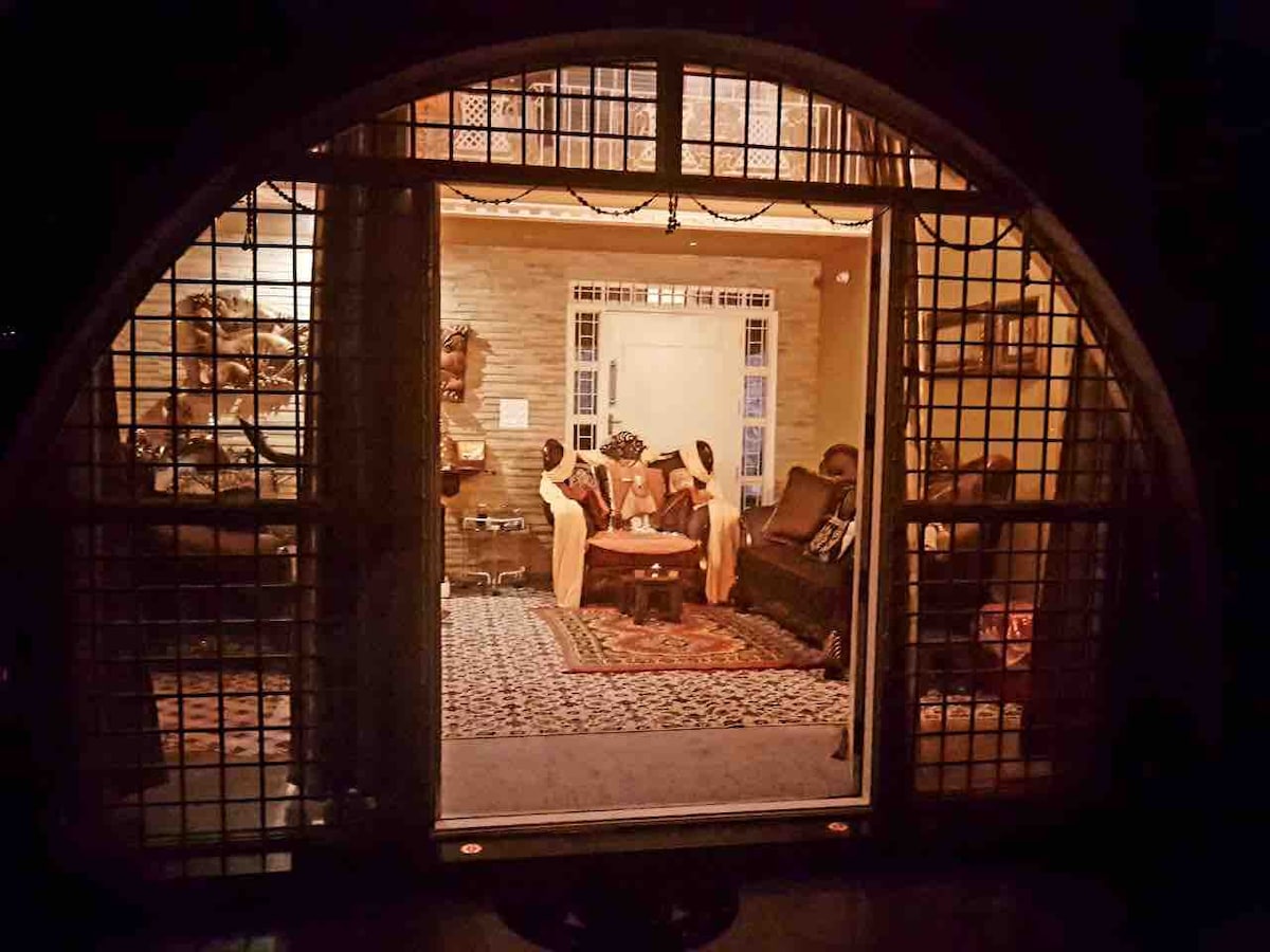 A warmly-lit living area is visible through a large arched doorway. The space features comfortable seating arranged around a low table, with a decorative rug covering the floor. Soft lighting creates an inviting atmosphere, highlighting the intricate details of the wall decor.