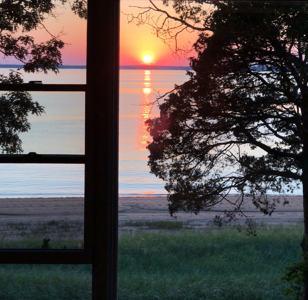 A vibrant sunset is visible through a window, casting a warm light over a calm bay. The sun reflects on the water, and silhouettes of trees frame the scene, creating a serene coastal atmosphere.
