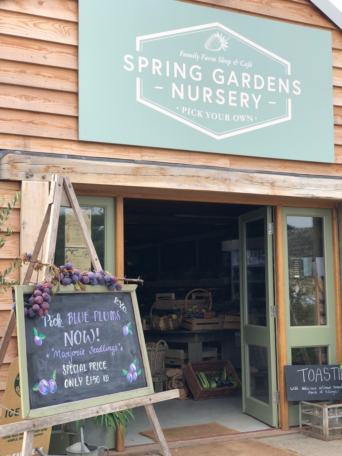 The entrance of a farm shop is displayed, featuring a wooden sign that advertises seasonal products. A welcoming open door reveals a glimpse of the interior, while a chalkboard highlights special offers for customers. The rustic exterior reflects the farm's natural surroundings.