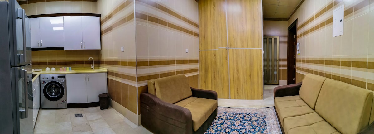 The image showcases a modern kitchen and living area featuring two sofas arranged around a patterned area rug. A washing machine and refrigerator are visible, alongside a countertop. The light-colored tiled walls and wooden accents create a welcoming environment.