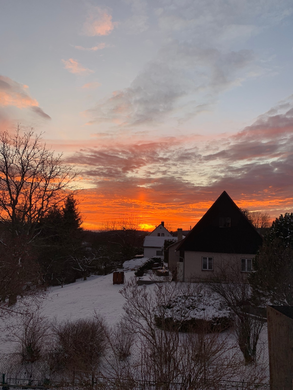 A sunset sky is filled with vibrant hues of orange and pink, reflecting beautifully over a snowy landscape. A simple house with a distinctive roof shape is visible along with tree silhouettes, adding depth to the tranquil scene.