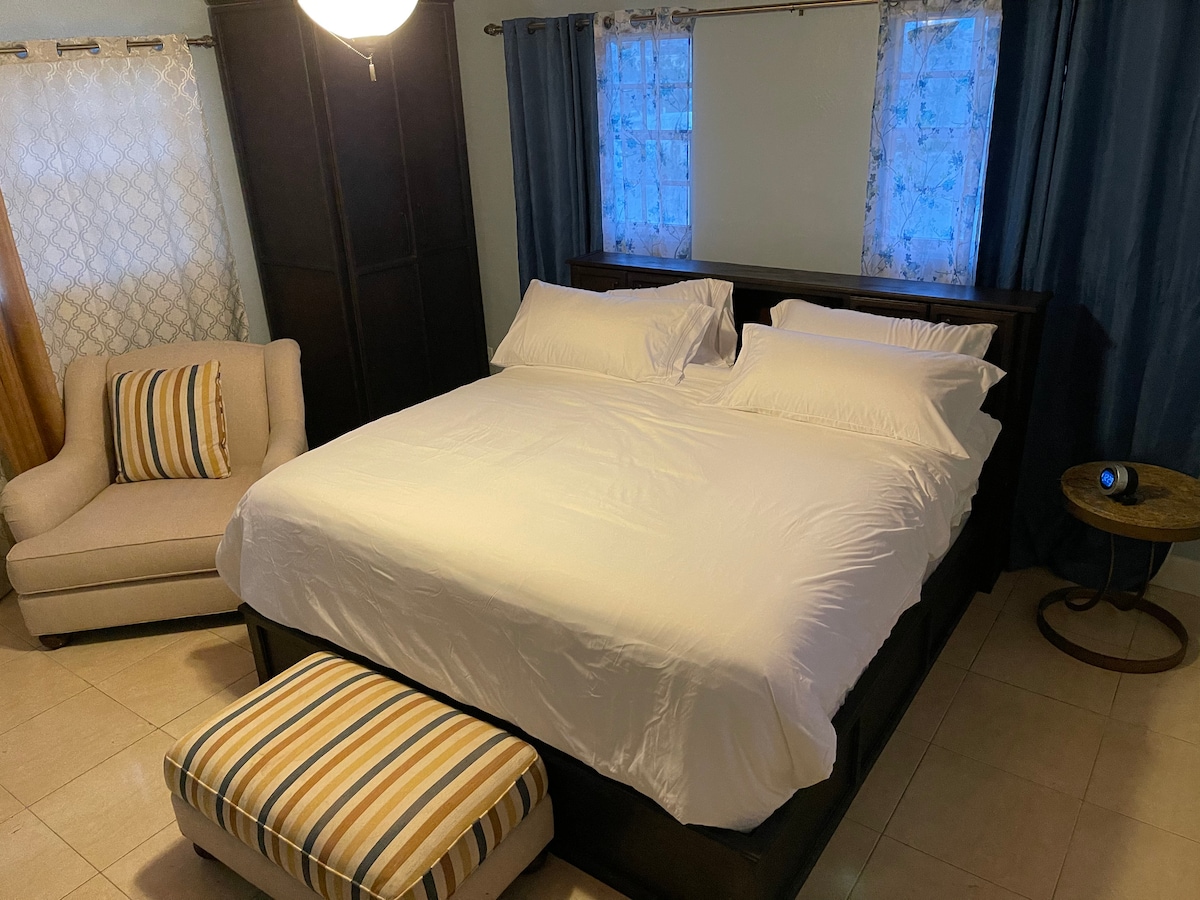 A spacious sleeping area is highlighted by a large bed with crisp white linens. A cozy armchair and a striped ottoman provide additional seating. Natural light enters through two windows adorned with sheer curtains, adding a soft glow to the room's neutral tones.