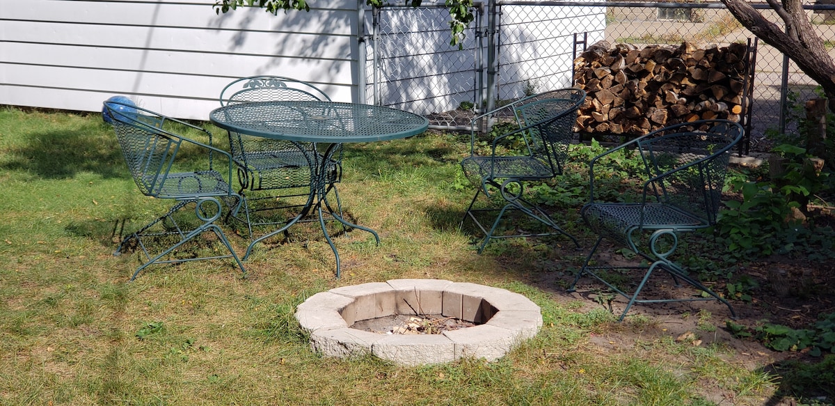 A green metal table and four matching chairs are arranged in a grassy area of the backyard. A stone fire pit is located nearby, surrounded by a tidy lawn. Wood is stacked against a fence in the background, contributing to the outdoor ambiance.