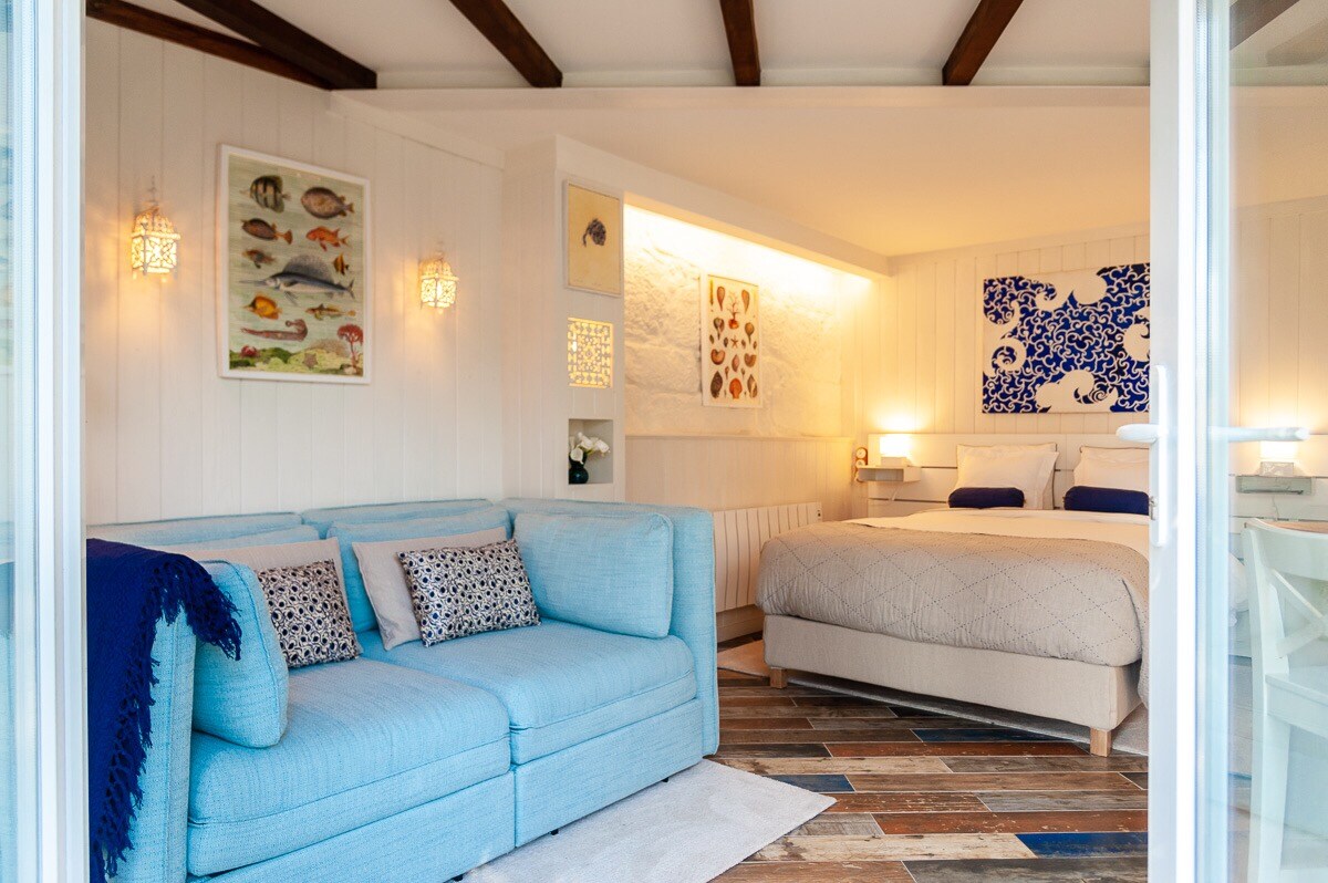 A light-filled studio features a spacious double bed with elegant bedding and a cozy blue sofa in the sitting area. Textured wooden flooring adds warmth, while artwork on the walls complements the maritime theme. Soft lighting creates a comfortable ambiance throughout the space.