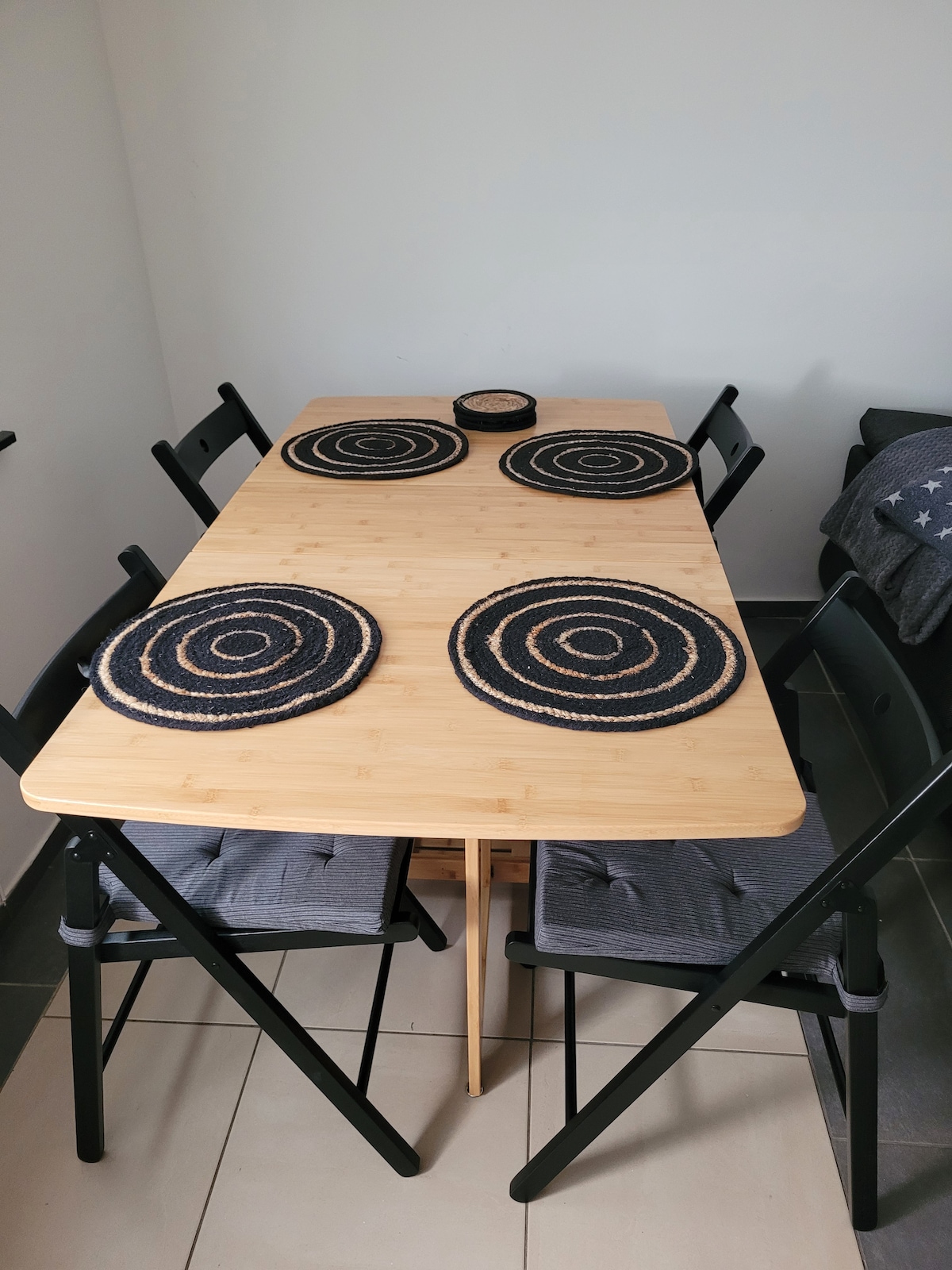 A simple dining table is equipped with four chairs, featuring grey cushions. Circular placemats in natural tones are arranged on the table's surface, which is made of bamboo. The clean lines and minimalistic design create a practical dining space.
