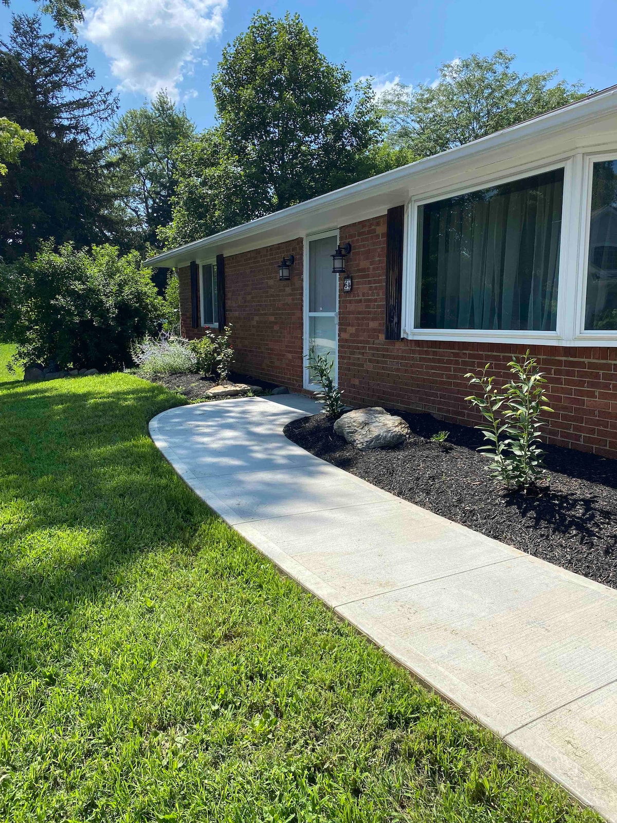 The exterior of the guest house is visible, showcasing a well-maintained pathway leading to a front door. Lush greenery surrounds the brick façade, complemented by landscaping features including small plants and a decorative rock. Bright natural light enhances the inviting entryway.
