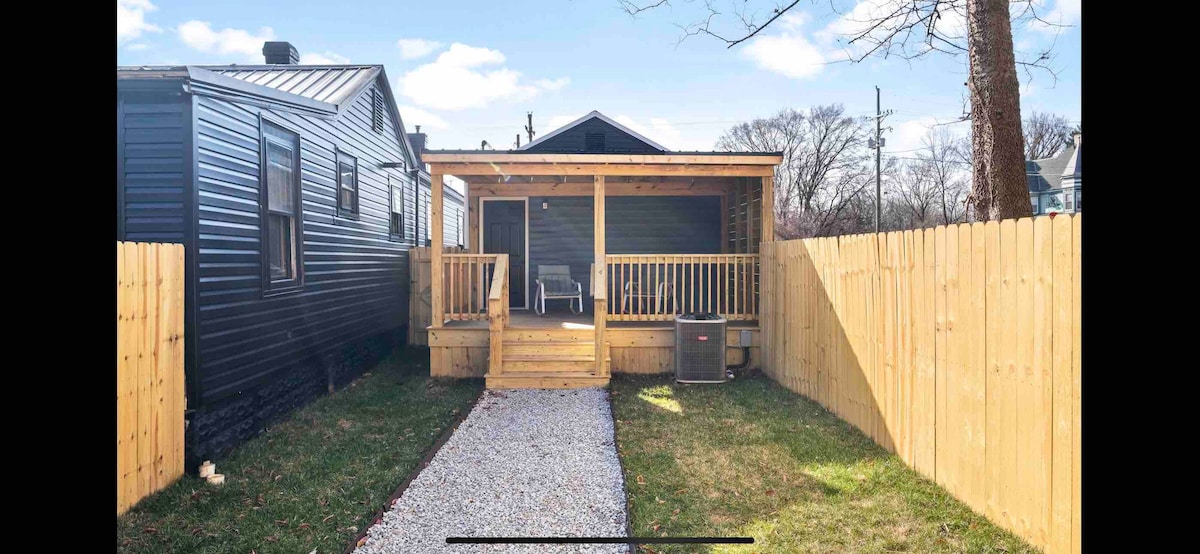 A covered porch is framed by a wooden railing, offering seating arrangements. The entrance features a gravel path leading to the porch, surrounded by a neatly maintained lawn and a fenced yard. Nearby, a dual-unit air conditioning unit is positioned discreetly.