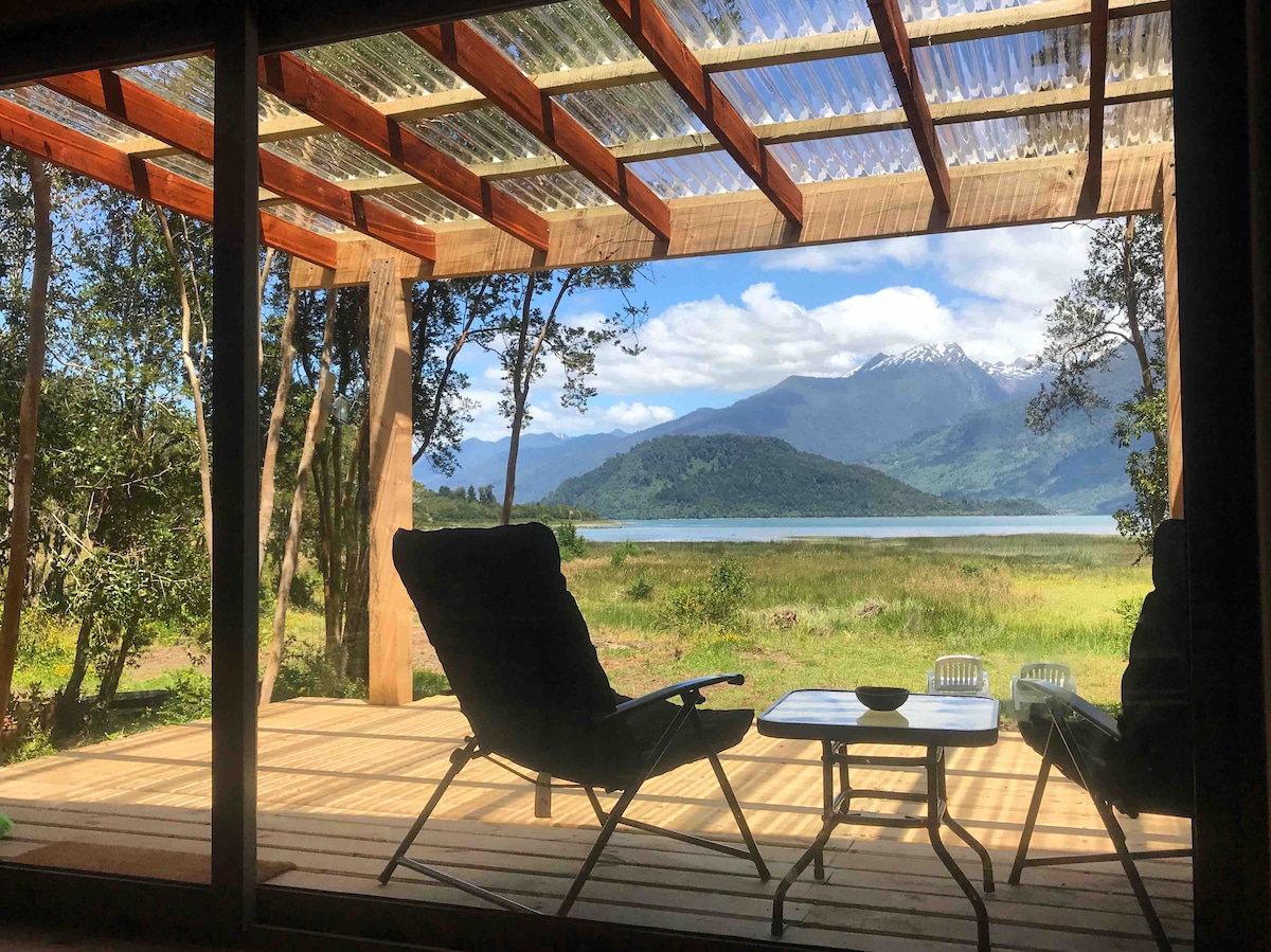 A covered outdoor area features two comfortable chairs and a small table, overlooking a serene landscape. Scenic views include a verdant shoreline and distant mountains, with large glass doors providing an inviting connection to nature.