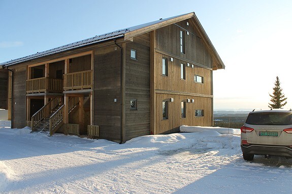 A two-story wooden building is depicted, featuring a sloped roof and multiple windows allowing natural light inside. The entrance includes a set of steps leading to a balcony. A snow-covered landscape surrounds the property, with a parked vehicle visible nearby.