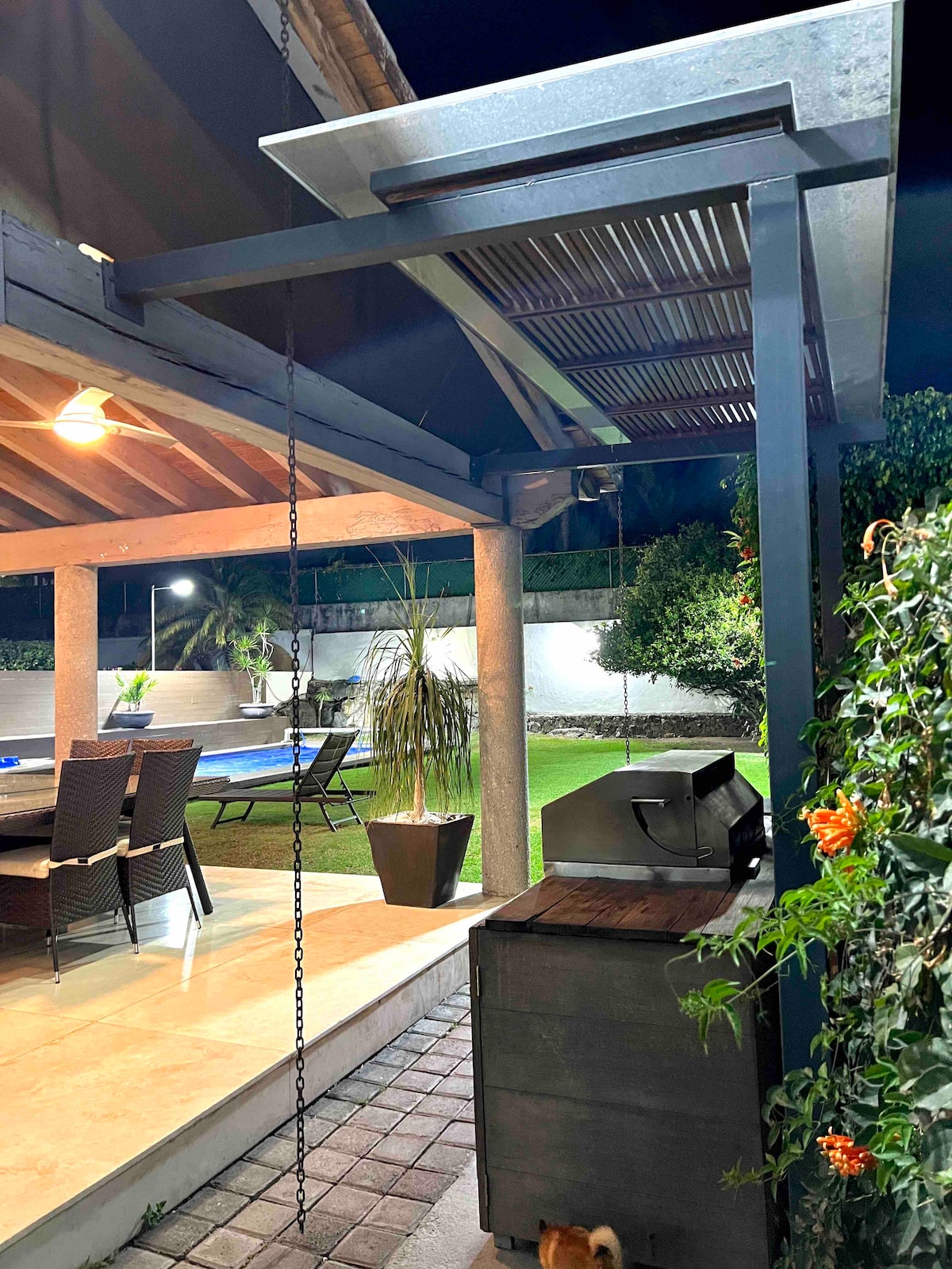 An outdoor terrace reveals a charming dining area under a shaded roof, featuring a wooden table surrounded by six comfortable chairs. A barbecue grill stands nearby, next to a small storage unit. Soft garden lighting creates a relaxed ambiance, enhancing the lush greenery in the background.