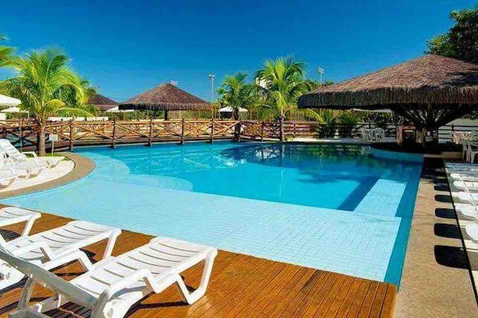 A large swimming pool is surrounded by wooden decking and lounge chairs. Thatched-roof cabanas provide shaded areas, while tropical palm trees create a lush backdrop under the clear blue sky.