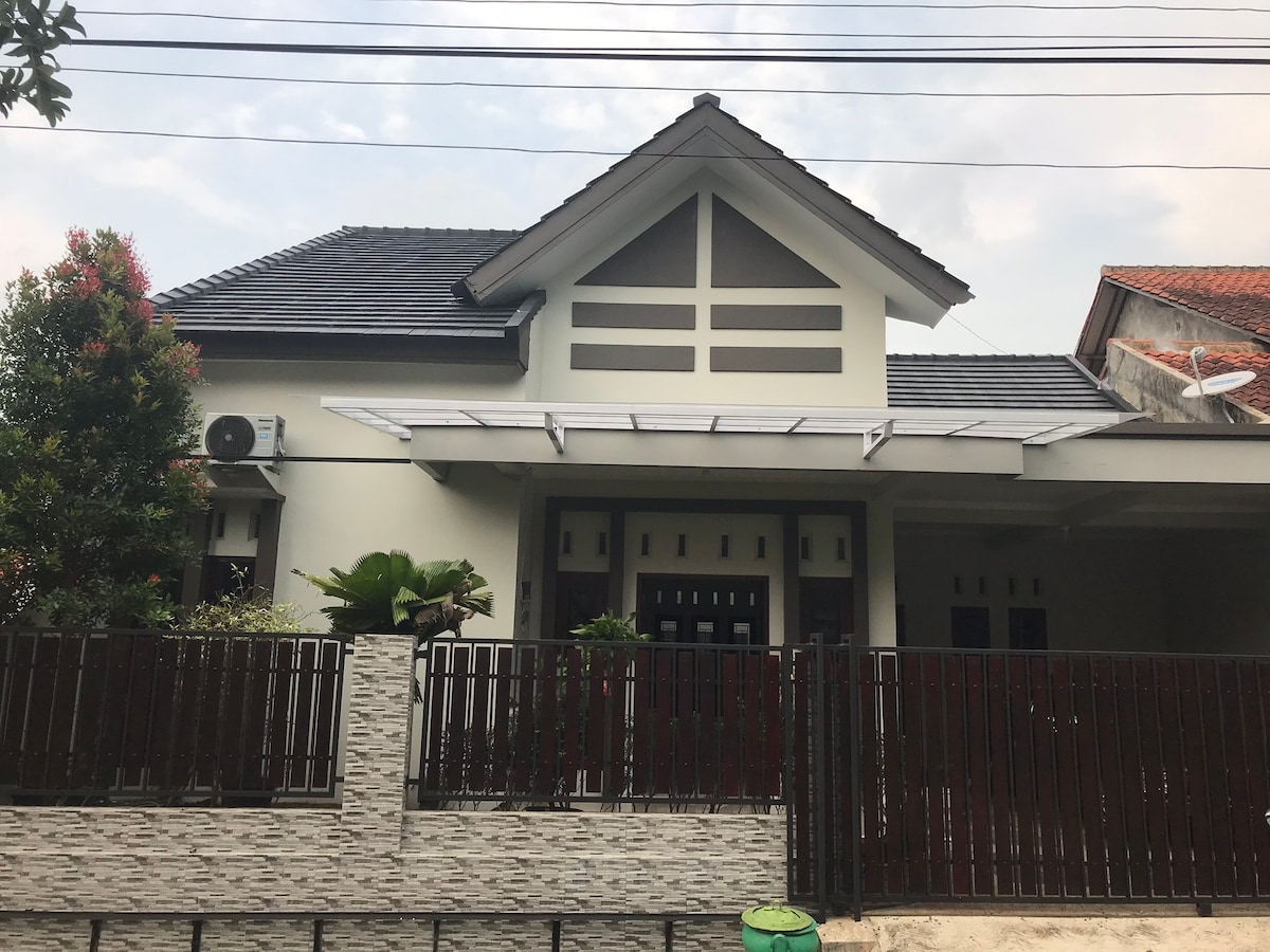 A modern house exterior is presented, featuring a peaked roof and a covered entryway. A combination of fresh greenery and a decorative fence enhances the facade. Large windows allow natural light to enter the space, with air conditioning units visible on the side.