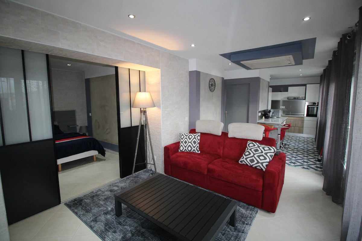 A modern living space is visible, featuring a red sofa with patterned cushions opposite a dark coffee table. A dining area with four chairs is seen to the right. Large sliding doors reveal a bedroom with a double bed. Soft lighting enhances the decor.