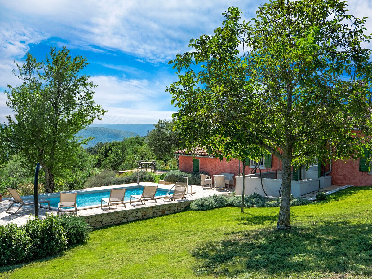 A self-standing house is set against a backdrop of rolling hills and clear blue skies. A private swimming pool is surrounded by sun loungers, with a spacious lawn and trees offering shade. Flowering plants are visible, enhancing the serene outdoor environment.