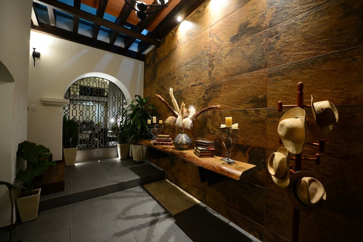 A hallway features a textured stone wall with natural lighting highlighting decorative elements. A wooden table displays decorative items, and potted plants add greenery. A coat rack with hats stands against the wall, and an arched doorway leads to the next area.