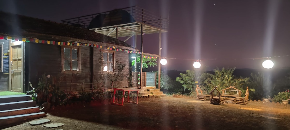 A rustic structure with wooden panels is set against a night sky. Soft lighting illuminates the area, highlighting a staircase leading to an upper deck. Colorful lanterns are suspended above an open space, creating a cozy atmosphere for gatherings amidst greenery.