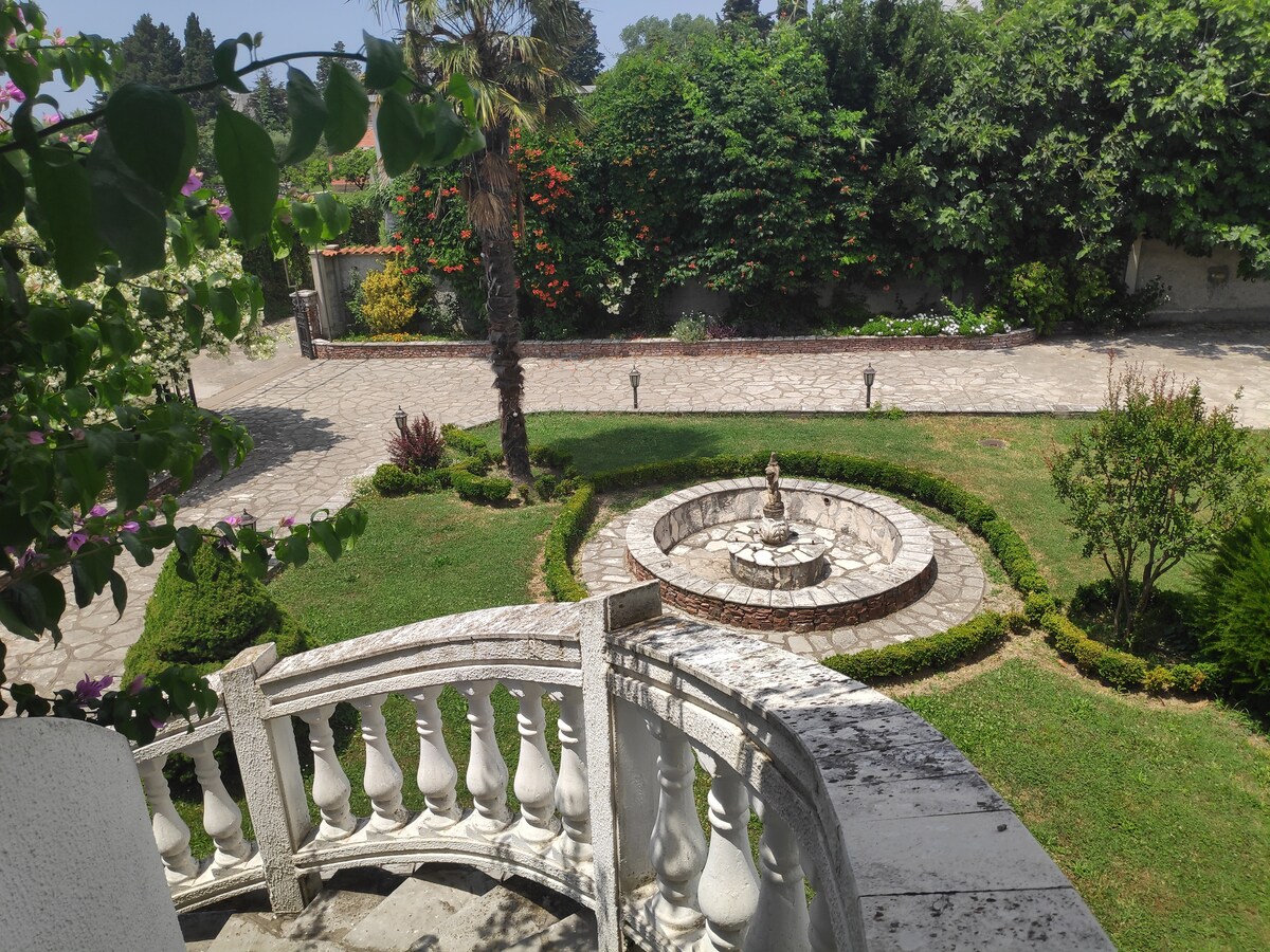A view from a balcony overlooks a well-maintained garden featuring a circular stone fountain at the center. Lush green hedges and blooming flowers surround the area, while pathways are lined with elegant light fixtures and manicured lawns, creating a serene outdoor space.