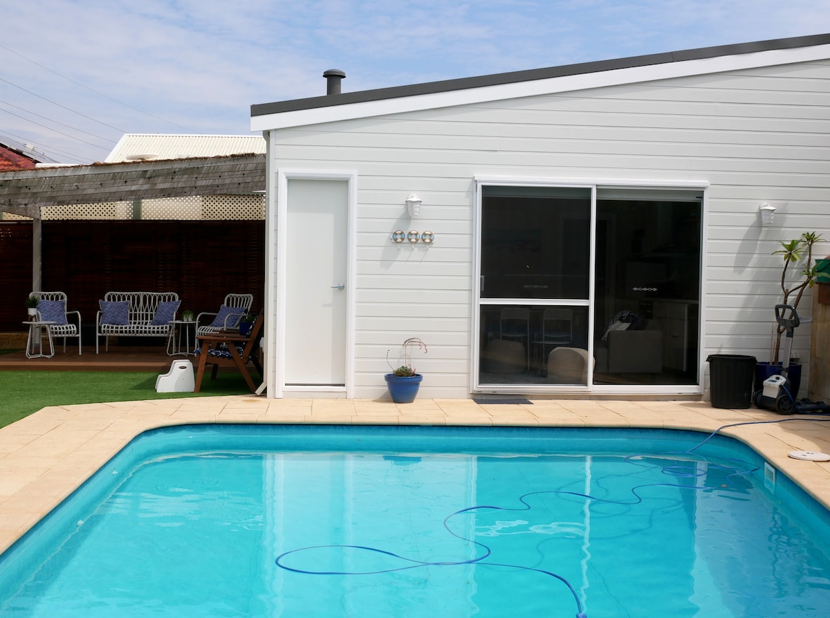 A bright pool area features a clear swimming pool surrounded by beige stone tiles. A light-colored house with large glass windows stands nearby, reflecting natural light. Outdoor seating is visible on the side, with a potted plant placed near the pool.