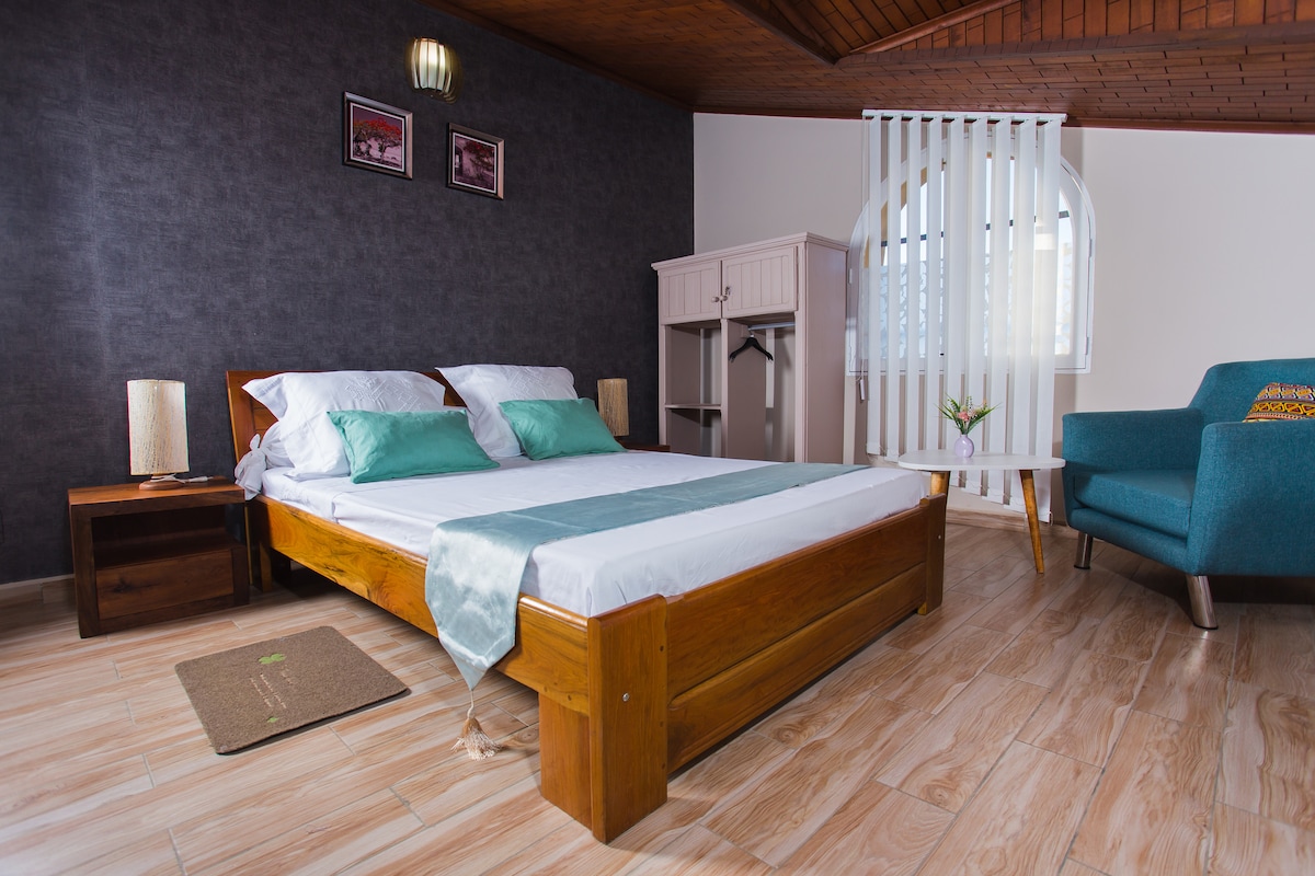 A spacious sleeping area is dominated by a wooden bed with crisp linens and decorative pillows. A light blue throw runs across the bed. A small nightstand with lamps is positioned beside the bed, and a wardrobe is visible in the background.