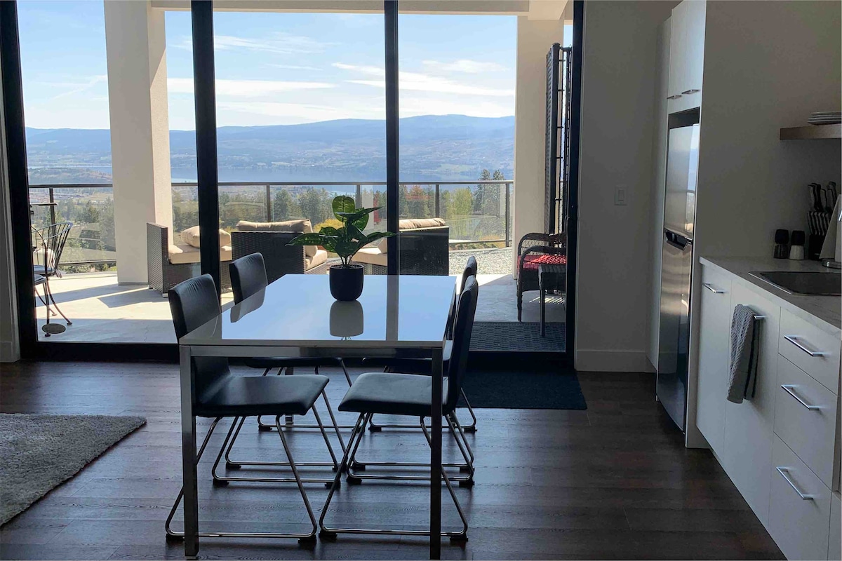 A dining area is visible, featuring a rectangular table surrounded by black chairs. Large glass doors provide a scenic view of Okanagan Lake and the surrounding landscape. The outdoor seating area can be seen through the open doors, enhancing the connection between indoor and outdoor spaces.
