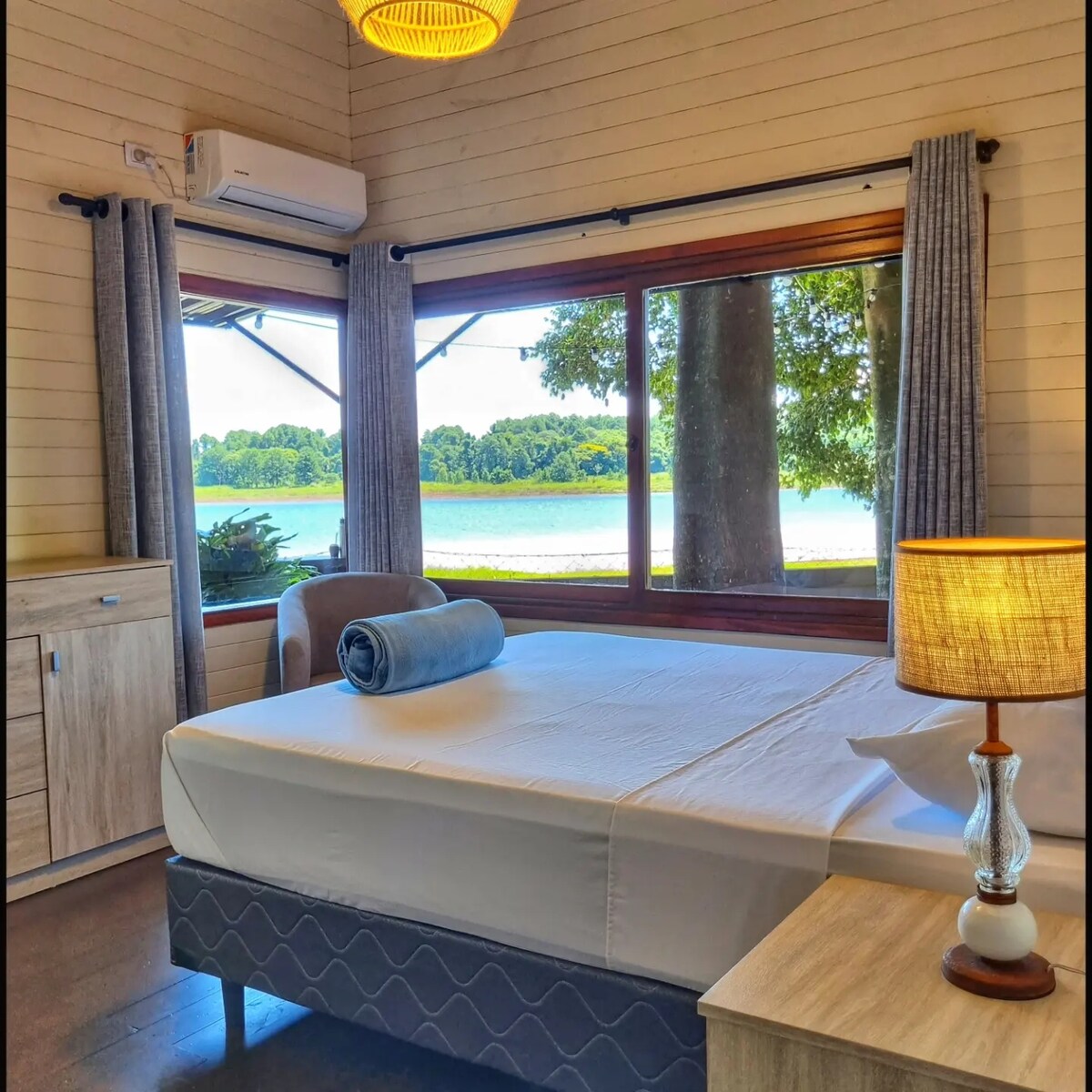 A spacious sleeping area features large windows that frame views of the lake and surrounding greenery. A neatly made bed is accompanied by a simple blue throw and a bedside lamp, while light curtains gently soften the natural light entering the room.