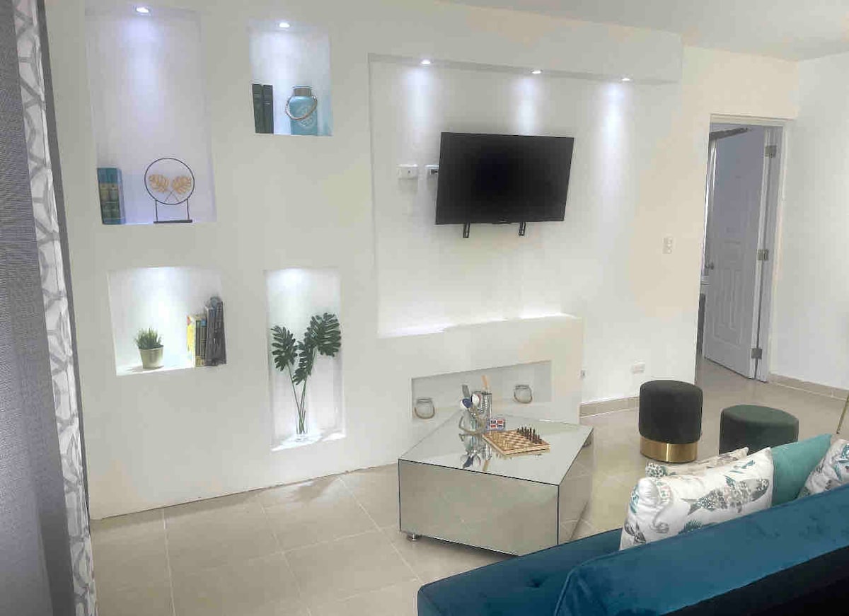 A modern living area features a stylish glass coffee table at its center, surrounded by a comfortable sofa. Wall-mounted shelves display various decorative items and plants. A flat-screen TV is mounted on the wall, with soft lighting enhancing the ambiance of the space.