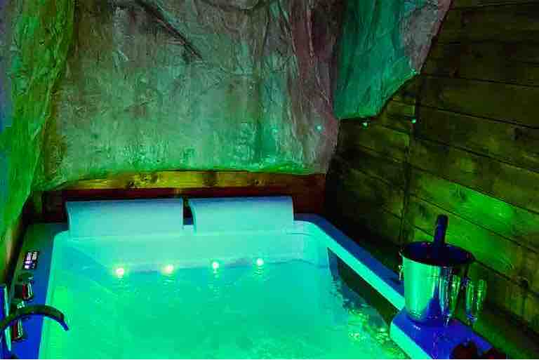 A mini hydro massage pool is situated in a cave-like setting, illuminated by soft green lights. The pool's water is calm, reflecting the colors above. A stylish ice bucket with champagne glasses rests nearby, enhancing the tranquil ambiance.