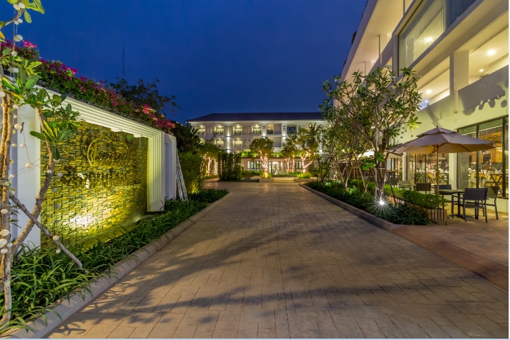 A tranquil pathway leads through lush greenery, lined by stone walls and flowering plants. Soft lighting casts a warm glow on the walkway, inviting guests toward the hotel structure visible in the background. Outdoor seating areas are elegantly arranged under umbrellas, enhancing the serene setting.