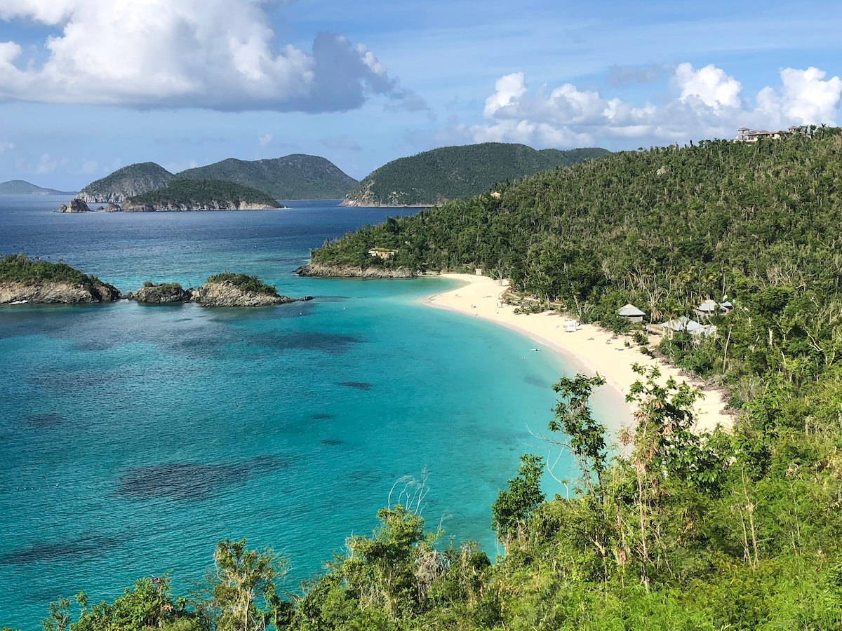 An expansive view of a secluded beach showcases soft white sand contrasting with the turquoise waters. Lush green hills frame the shoreline, extending to several small islands in the distance. The sky is partly cloudy, enhancing the serene coastal landscape.