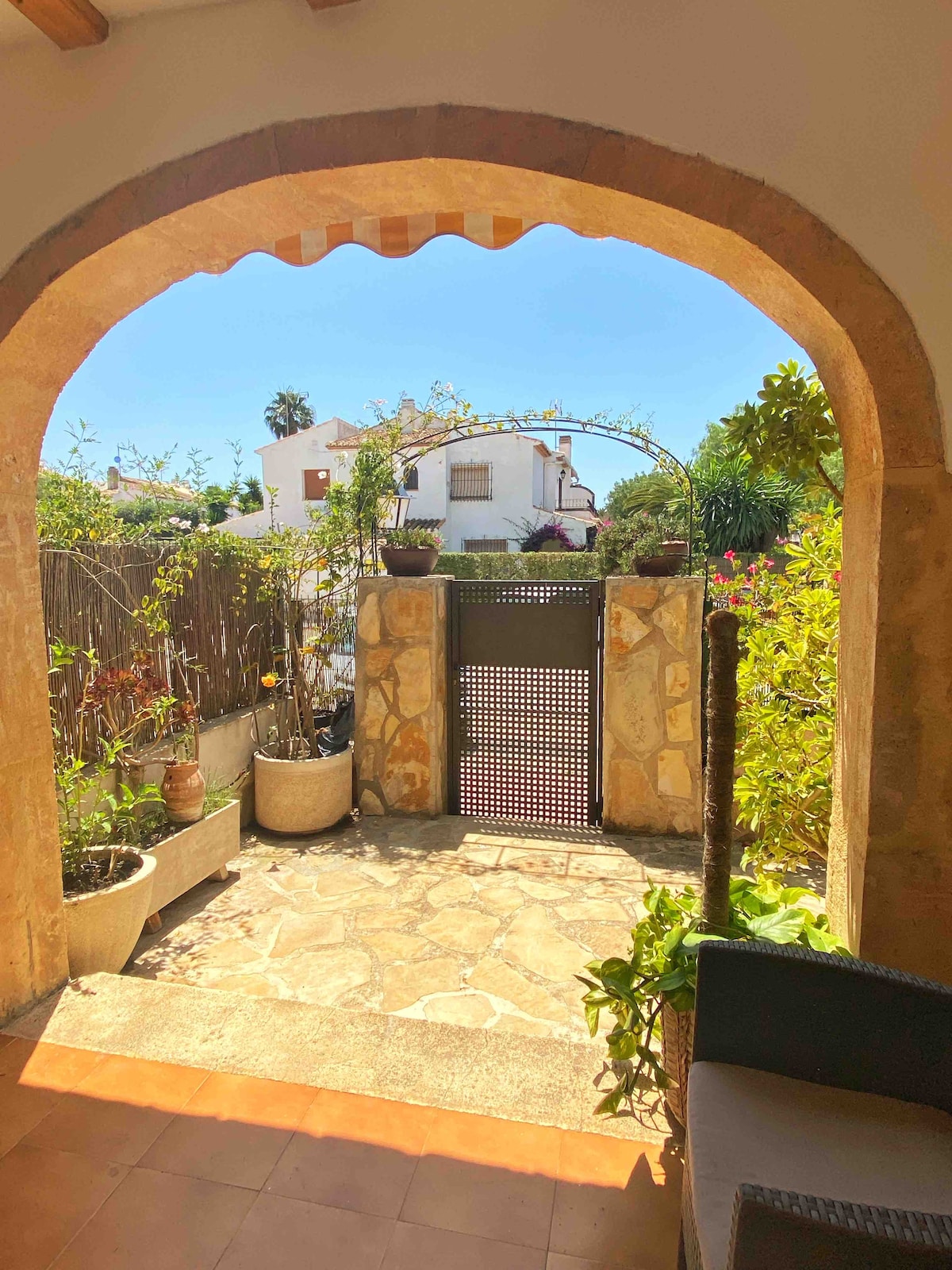 An outdoor entryway is framed by an arched stone doorway, leading to a garden visible beyond. Potted plants line the path, with a stone surface underfoot. A gate is seen in the distance, opening to a serene view of greenery and neighboring buildings.