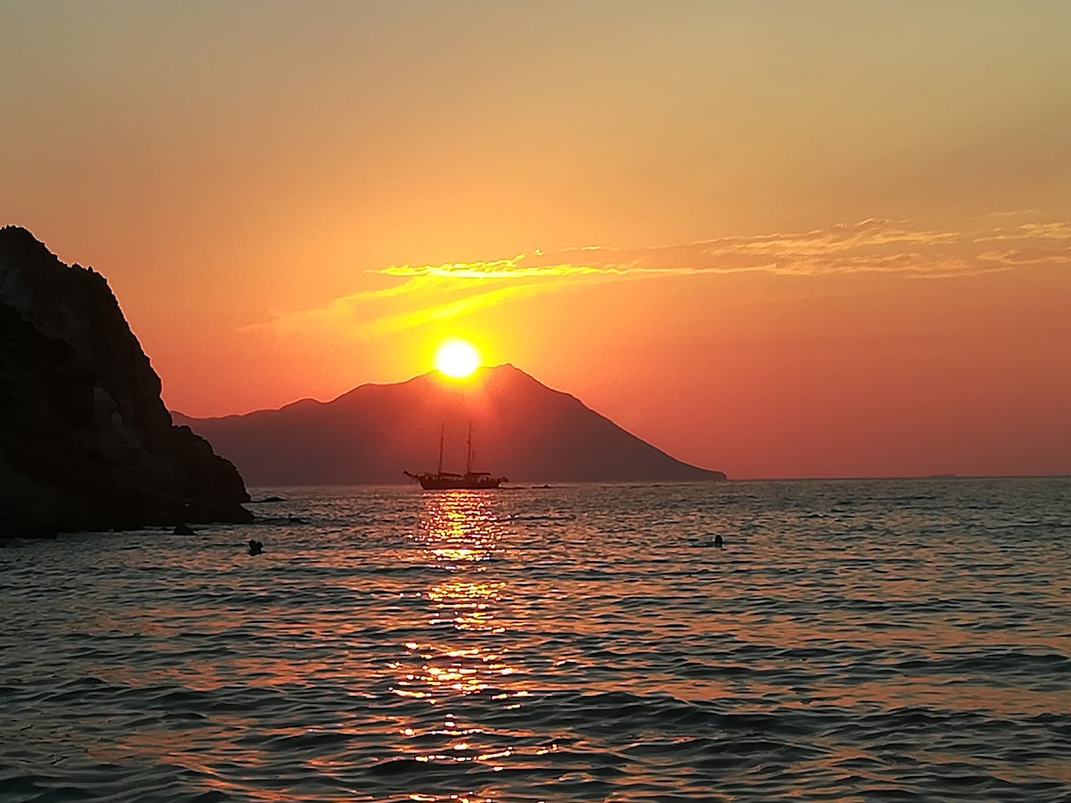 A serene seascape is captured at sunset, with golden rays reflecting off the water's surface. A silhouette of a sailboat is positioned against the backdrop of a mountainous shoreline, where the sun gently descends towards the horizon, casting warm colors across the sky.