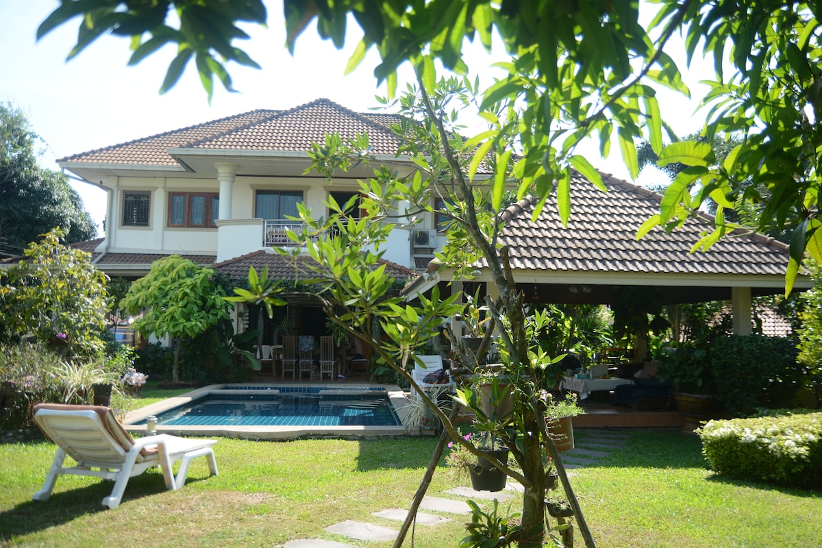 The two-story house is framed by lush greenery and a manicured lawn. A swimming pool is positioned in front, while a shaded seating area is visible under the patio. The roof features a traditional design, and vibrant plants surround the property, enhancing the outdoor space.