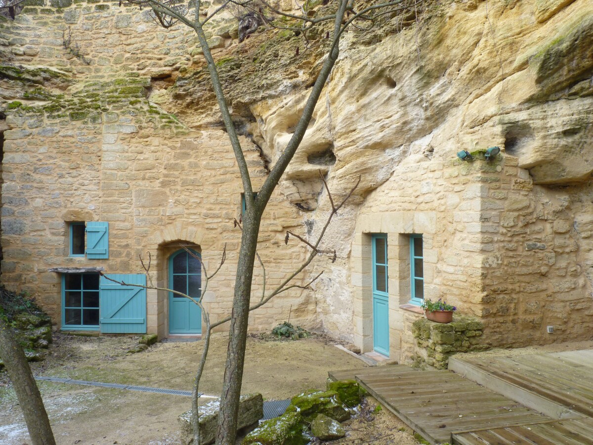 A stone exterior showcases a charming troglodyte dwelling, characterized by its warm beige tones and teal wooden doors and windows. The surrounding area features natural rock formations and a small, maintained pathway leading to the entrance. A tree adds a touch of greenery to the scene.