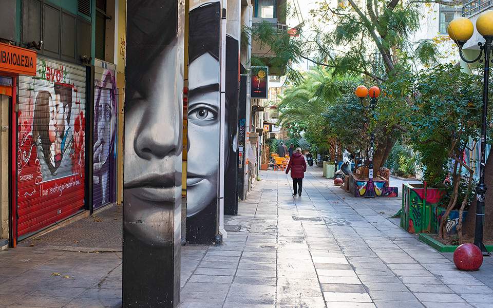 A quiet pedestrian street is lined with trees and colorful decorations. Small shops and vibrant murals can be seen on the walls. People stroll leisurely along the pathway, while outdoor seating areas with greenery create a relaxed atmosphere.