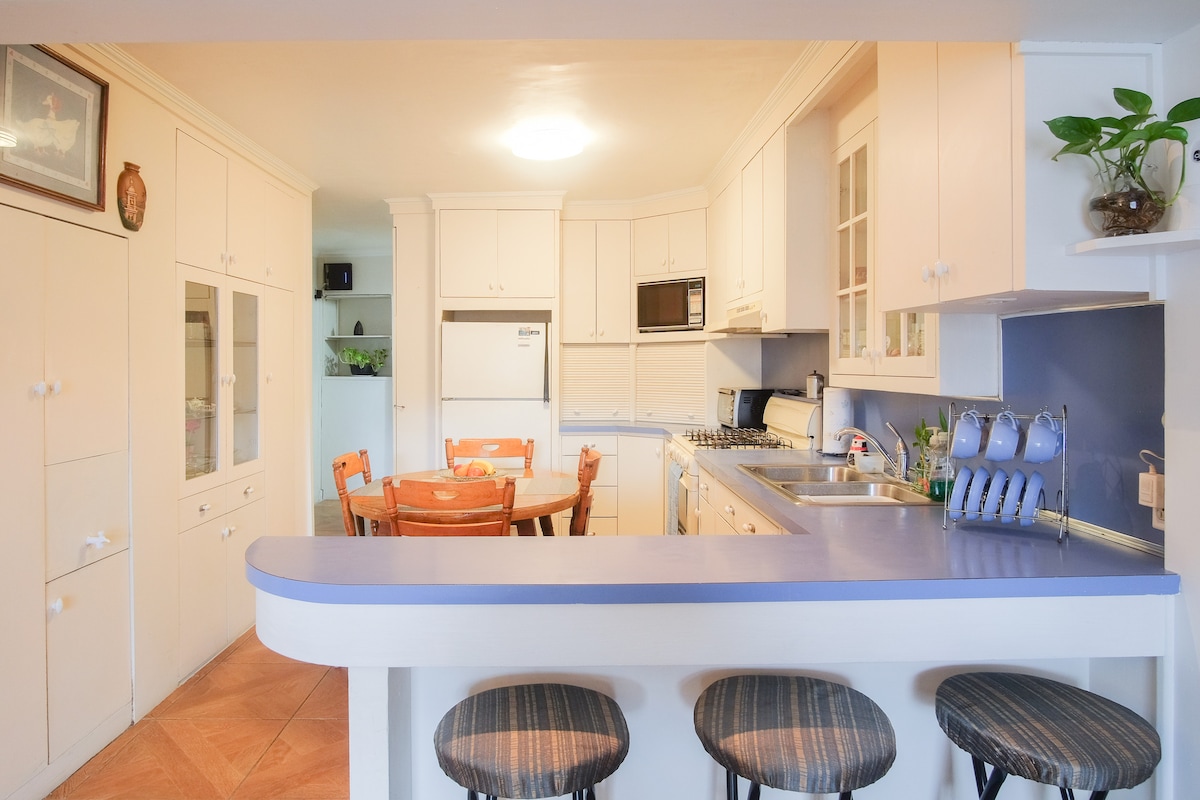 The kitchen features light-colored cabinetry and a double sink. A breakfast bar separates the kitchen from the dining area, which includes a wooden table surrounded by four chairs. Nearby appliances include a microwave and a refrigerator. Natural light fills the space.