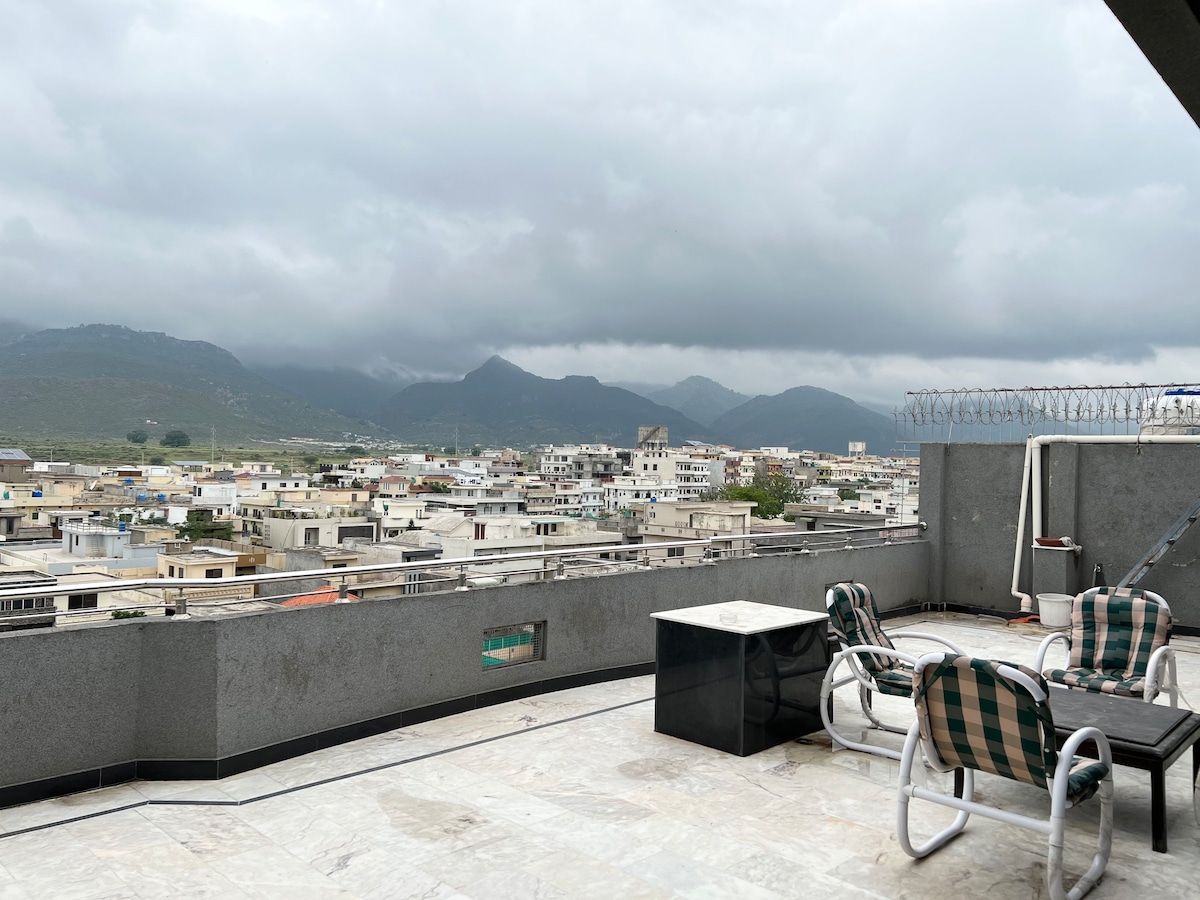 A spacious terrace offers panoramic views of the surrounding city and distant mountains. The area is furnished with a table and chairs, providing an inviting spot to relax outdoors. The surface is tiled, enhancing the open atmosphere.