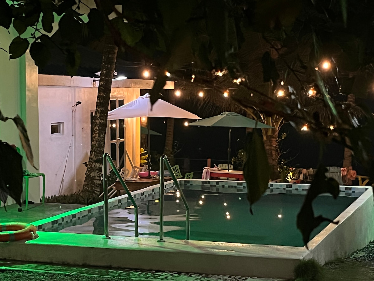 A swimming pool is illuminated by soft green lighting, reflecting the surrounding palm trees. Several lounge chairs are positioned nearby, complemented by umbrellas for shade. A portion of the villa can be seen in the background, along with strings of warm lights adding a relaxing ambiance.